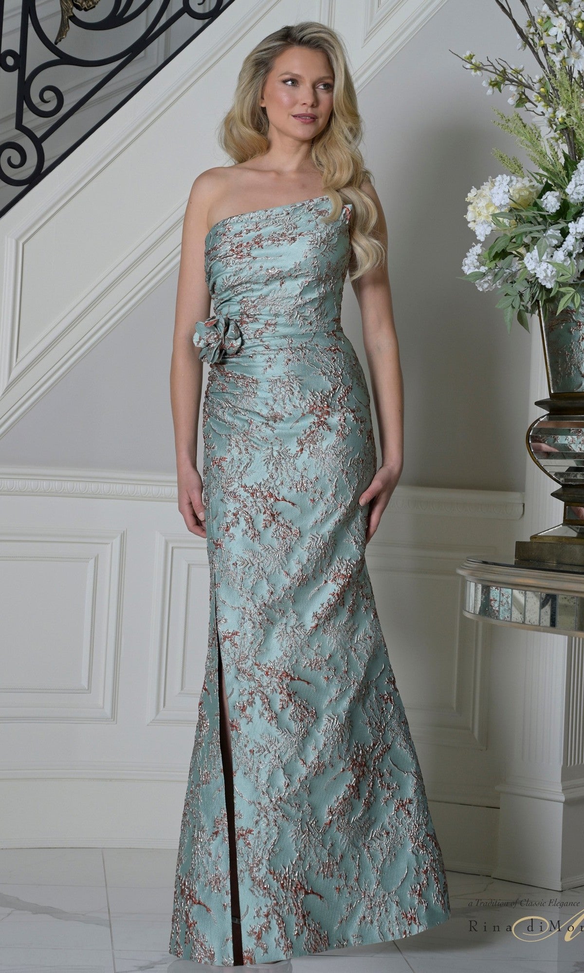 A woman wearing the Long Formal Dress: Rina Di Montella RD3502 in Sage Crimson stands indoors near a staircase and floral arrangement.